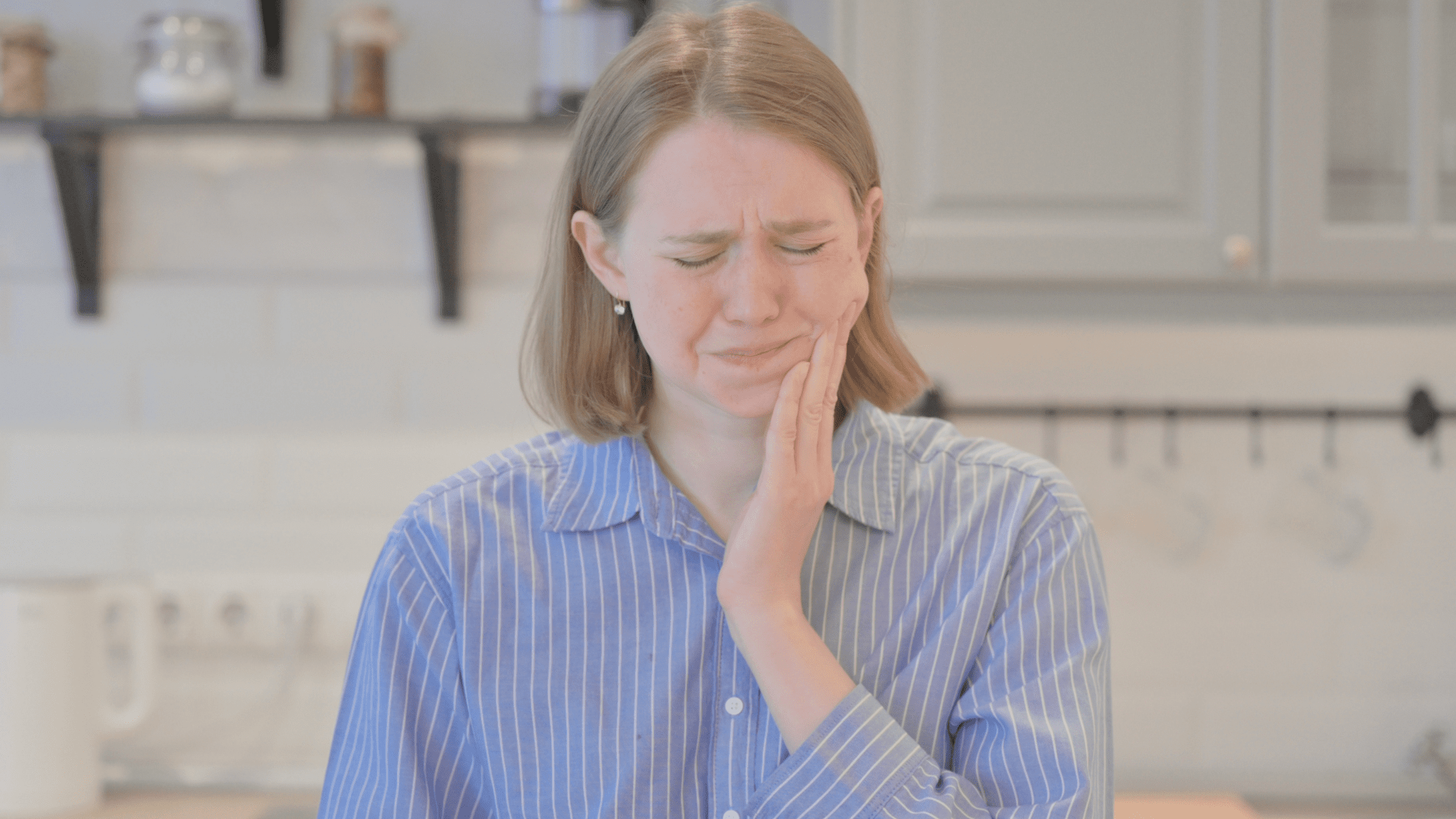 Woman standing in a kitchen, holding her cheek because she is experiencing a toothache.